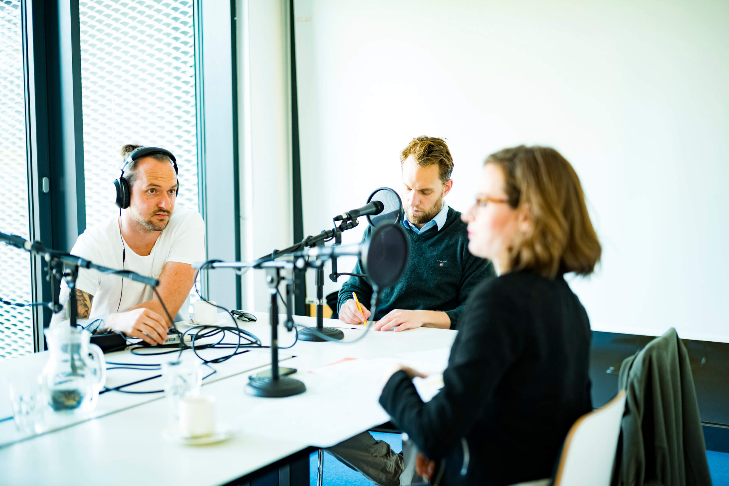 Podcast-Situation; 3 Personen sitzen vor Mikrofonen und nehmen einen Podcast auf